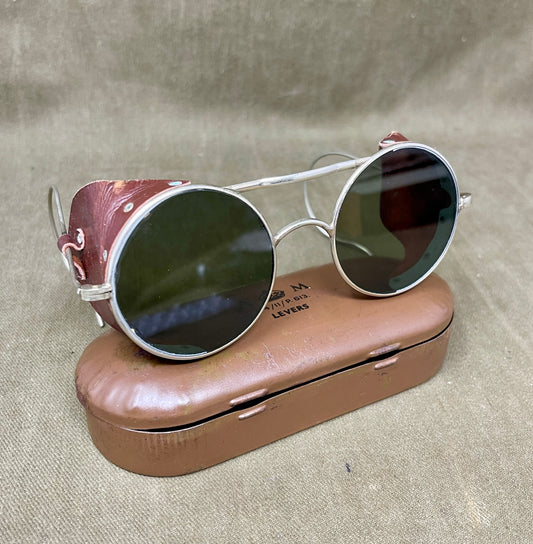 Round sunglasses with silver frame and dark lenses on a brown case against a beige background